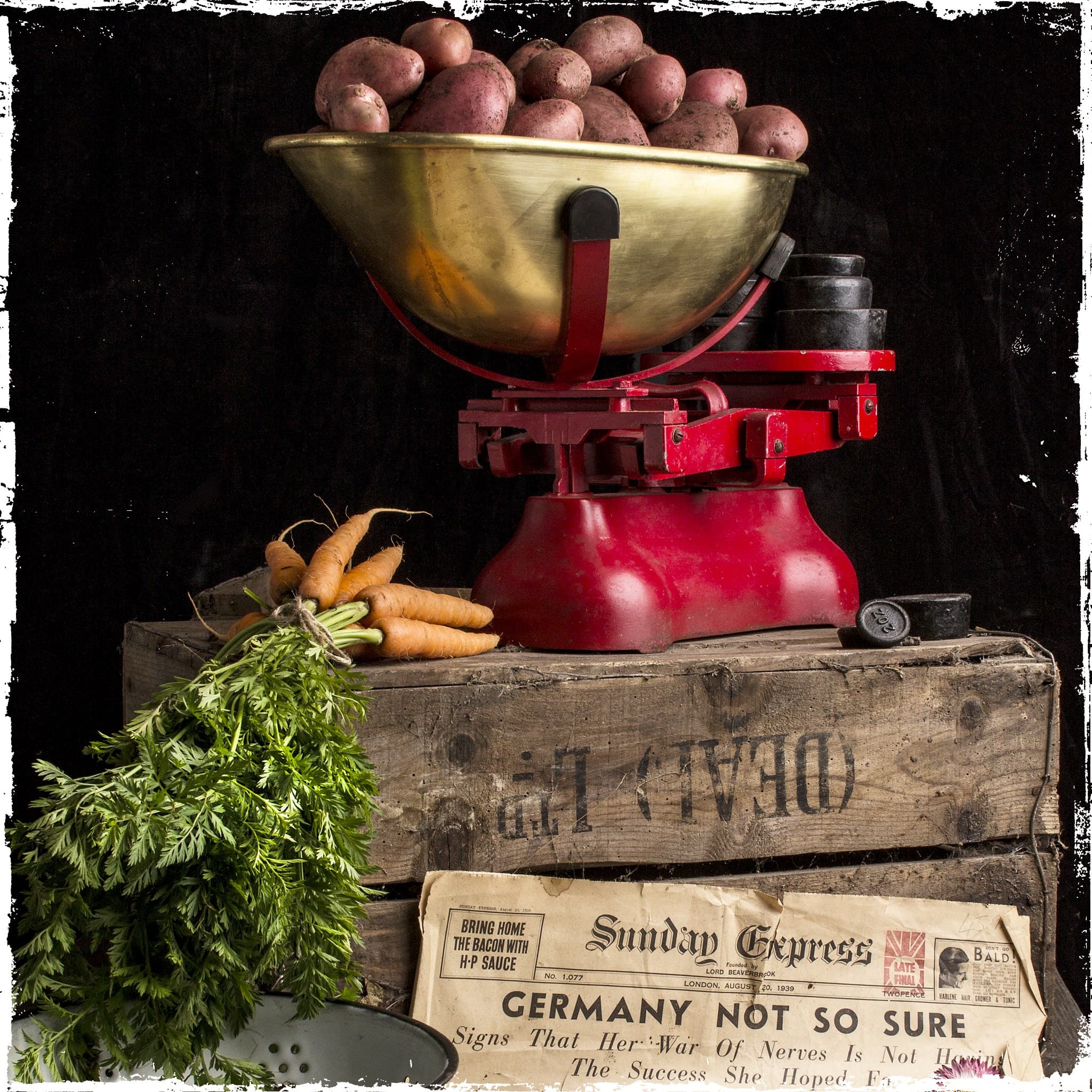 Vintage red grocer's scales with potatoes and carrots, on a wooden crate above a 1939 newspaper headlined 'Germany Not So Sure'